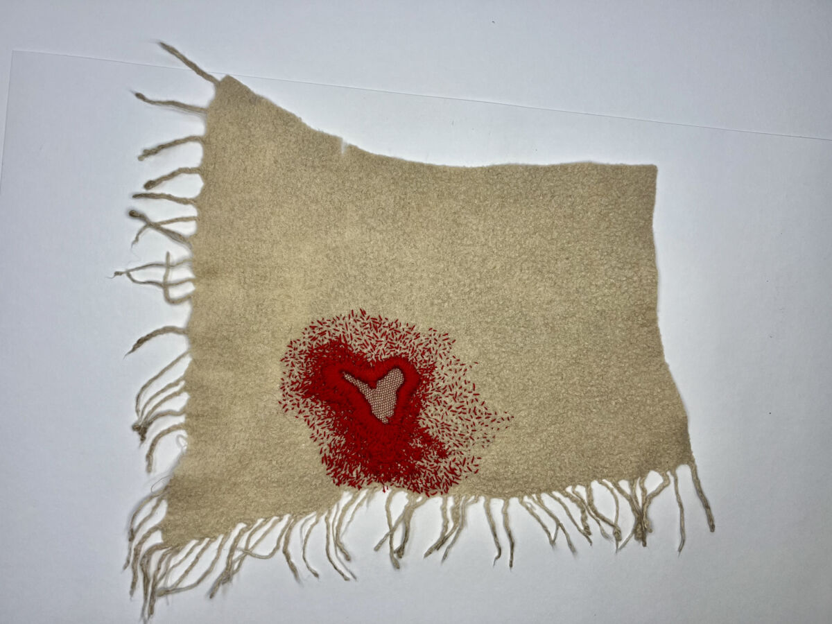 cloth with frayed edges, a red heart-shaped shape thickly embroidered fills the bottom half of the piece. It is surrounded by many small stitches and give the effect of bleeding